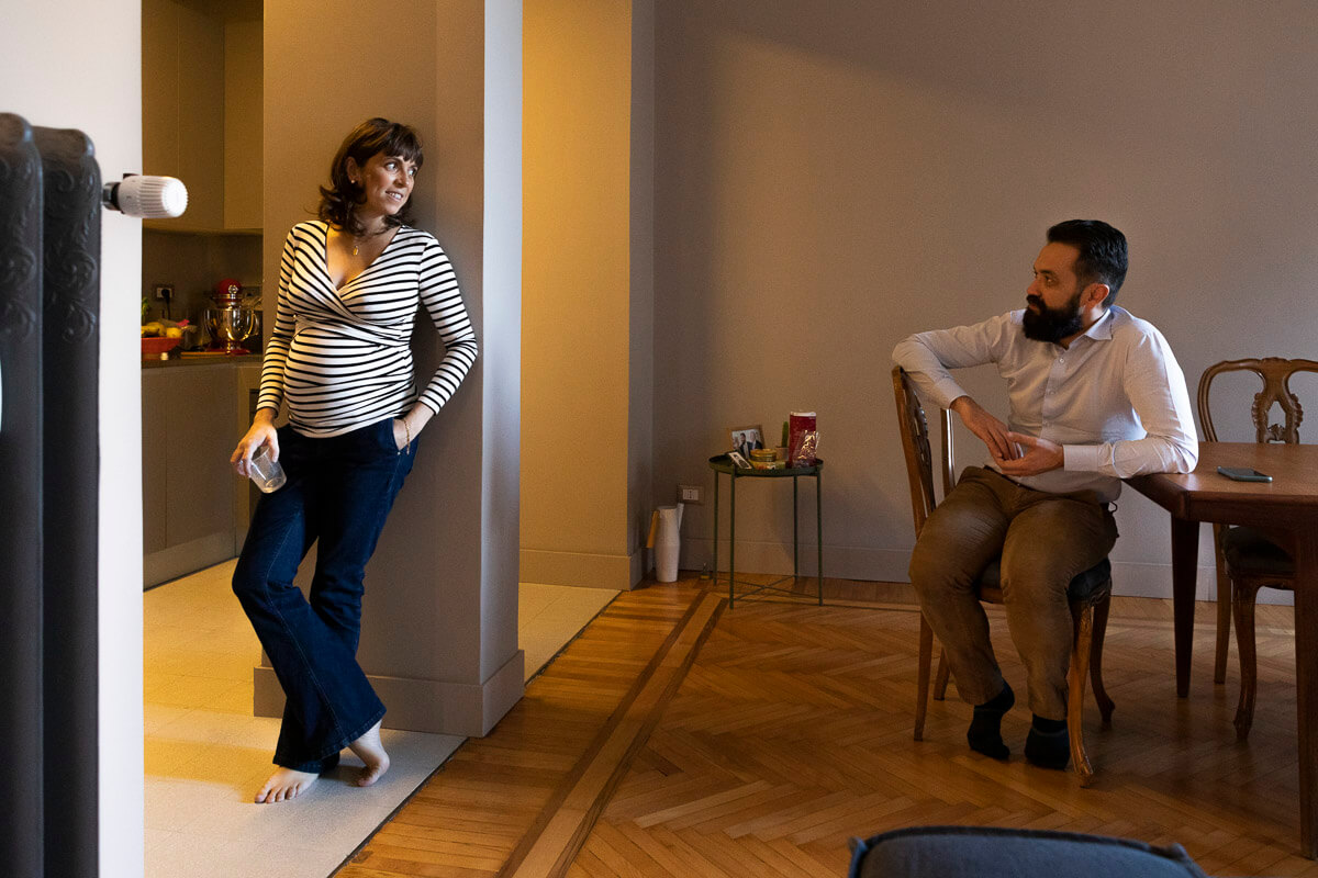 futuri mamma appogiata alla colonna in casa rivolta verso futuro papà seduto sulla sedia in salotto