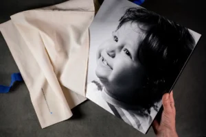 A person holding a large black-and-white portrait of a smiling child, partially removed from a protective fabric pouch. A person holding a large black-and-white portrait on fineart paper of a smiling child, partially removed from a protective fabric pouch.
