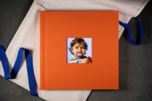 An orange photo book with a square cut-out cover featuring a smiling child. An orange portrait photo book with a square cut-out cover featuring a smiling child.