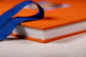 A close-up of an orange hardcover book with a blue ribbon bookmark. A close-up of an orange hardcover family album with a blue ribbon
