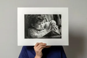 A person holding a black-and-white photo of a young child reading. A person holding a black-and-white fine art print in passpartout of a young child reading.