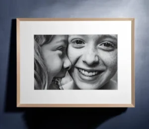 A framed black-and-white photograph of two smiling children, with one whispering to the other. A framed black-and-white fine art print photograph of two smiling children, with one whispering to the other.