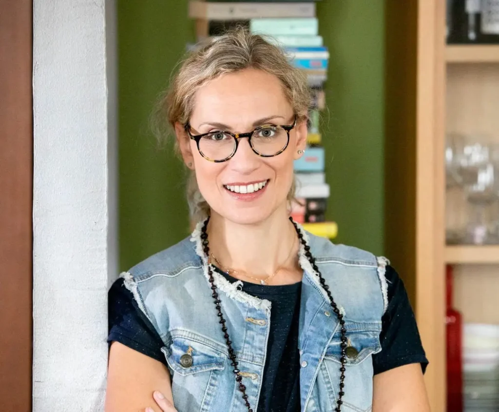 A smiling woman wearing glasses and a denim vest, standing in front of a bookshelf.
