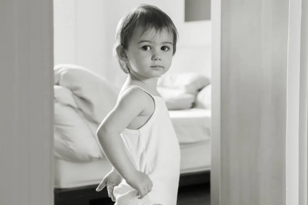 A black-and-white image of a toddler in a white outfit, standing confidently in a bedroom.