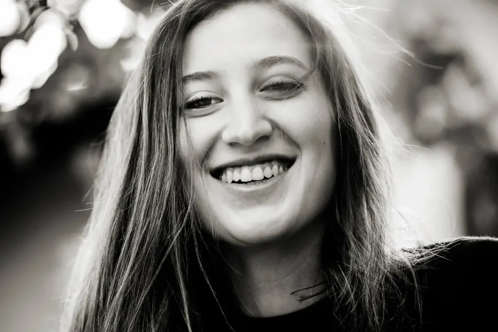 A black-and-white close-up of a young woman smiling brightly with slightly tousled hair.