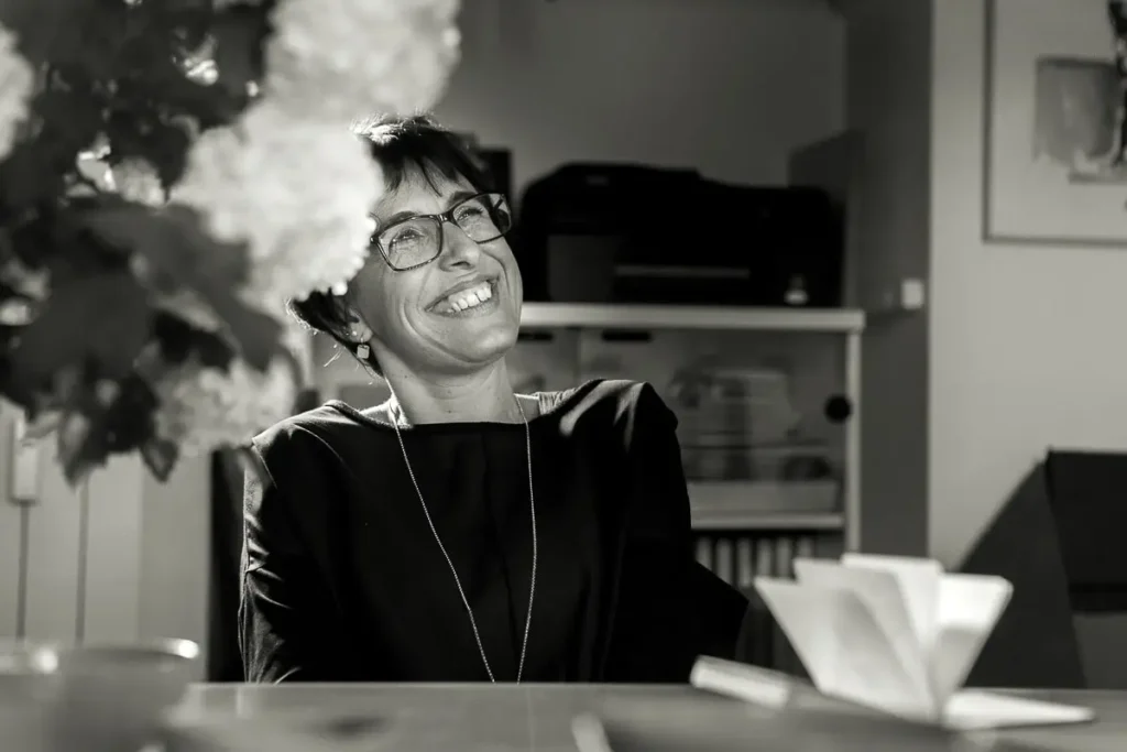 A black and white photo of a woman wearing glasses, smiling while sitting at a desk with a blurred foreground of flowers.