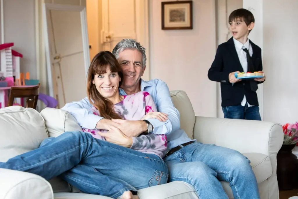 A happy couple sitting on a couch, embracing while their son walks by with a tray.