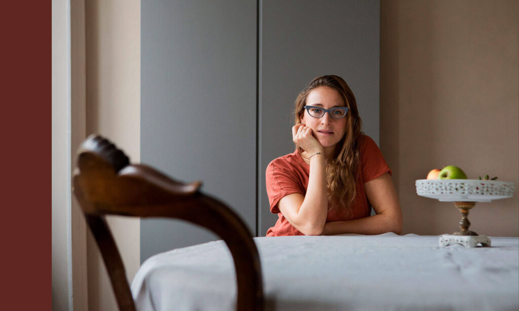 A woman with glasses and long wavy hair sitting at a dining table, resting her face on her hand.