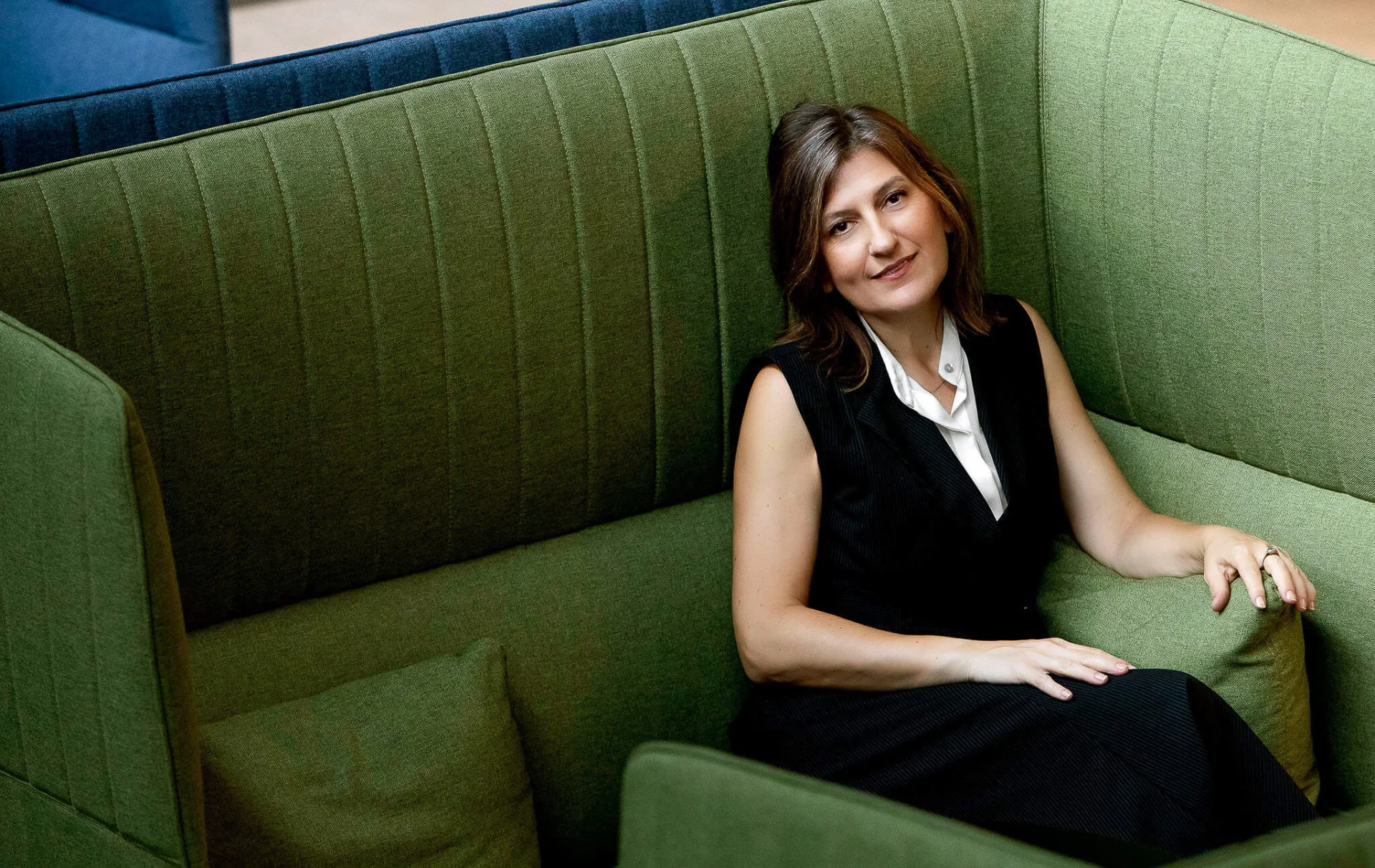 A woman in a black sleeveless outfit reclining on a green cushioned chair, smiling at the camera.