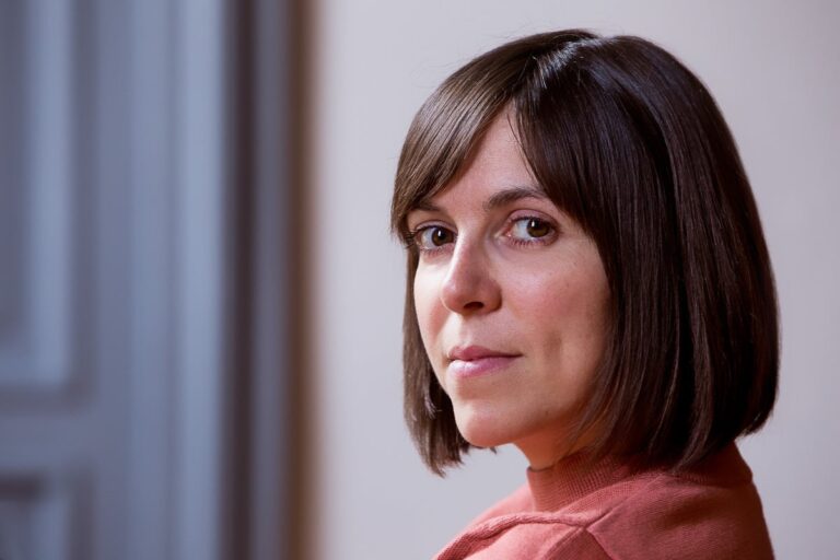 headshot of woman with orange turtleneck with gray door blurred in background