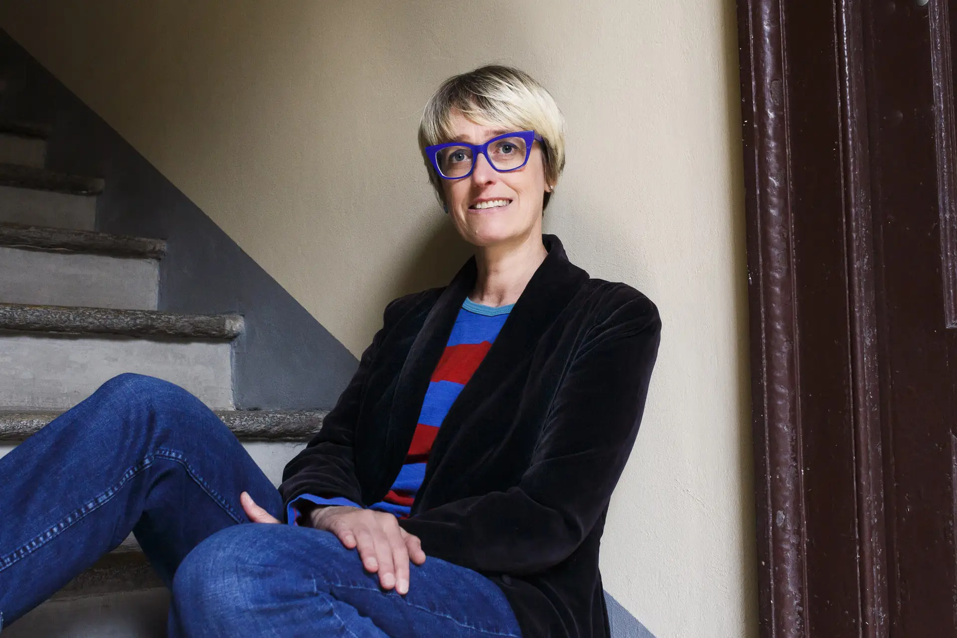 Una persona con capelli corti e biondi e occhiali blu siede sulle scale, sorridendo. Indossa una giacca nera, jeans blu e una camicia a righe blu e rosse, la scena perfetta per la storia di una fotografa professionista, con una parete neutra e una porta marrone scuro alle spalle.