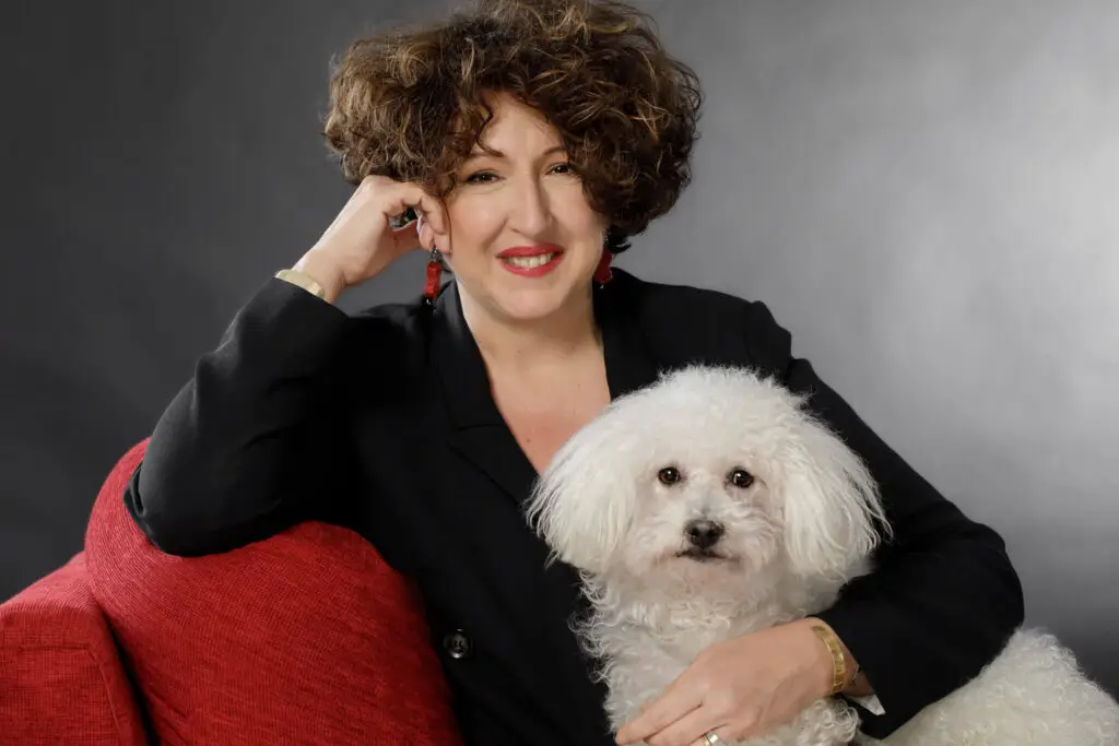 A woman in a black suit sitting confidently on a red couch with a fluffy white dog.